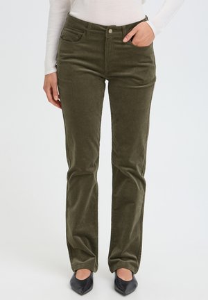 Personne portant un pantalon droit vert olive, un haut blanc à manches longues et des chaussures noires pointues, avec une main dans la poche.