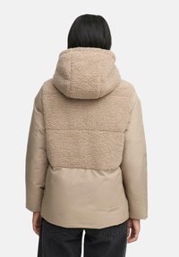 Beige pufferjack met een fluffy tan fleece achterkant en capuchon, met een gladde, glanzende voorkant en een merketiket aan de zoom.