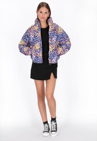Barevná zkrácená péřová bunda s mnohobarevným leopardím potiskem, sezipovým uzávěrem a elastickými manžetami. Nositá přes černý top a sukni.
