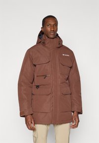 Veste isolée marron avec capuche, plusieurs poches sur le devant, fermetures éclair et boutons-pression, et un logo sur la poitrine. Tissu texturé avec poignets réglables.