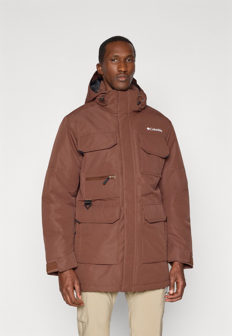 Veste isolée marron avec capuche, plusieurs poches sur le devant, fermetures éclair et boutons-pression, et un logo sur la poitrine. Tissu texturé avec poignets réglables.