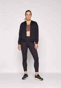Black zip-up hoodie over a burgundy sports bra, paired with black leggings and black athletic shoes. Smooth textures and fitted design.