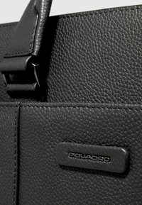 Borsa in pelle nera strutturata con accento hardware in argento visibile con il logo "Piquadro", che mostra dettagli di cucitura e un design elegante.