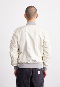 Giacca bomber di colore crema chiaro con colletto e polsini grigi a coste, caratterizzata da una texture liscia e lucida e tasche laterali. Indossata sopra pantaloni cargo scuri.