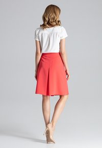 Koraalrode geplooide rok met een aansluitende taille, gecombineerd met een witte top. Het model draagt naaldhakken in huidskleur, wat een elegante silhouet van achteren laat zien.