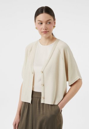 Beżowy sweter typu cropped z krótkimi rękawami, z zapięciem na guziki i subtelnymi detalami w formie oczek. Noszony na jasnym topie.