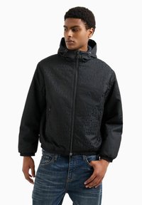 Giacca nera con cappuccio, caratterizzata da un motivo logo sottile, chiusura con zip e polsini elasticizzati. Abbinata a jeans di denim blu.