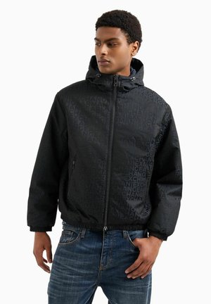Giacca nera con cappuccio, caratterizzata da un motivo logo sottile, chiusura con zip e polsini elasticizzati. Abbinata a jeans di denim blu.