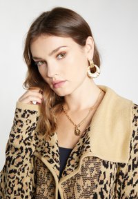 Beige Pullover mit Leopardenmuster, mit einem Schalkragen. Ein kräftige goldene Kettenhalskette und große Ohrringe runden den Look ab. Glatte Texturen.