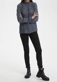 Grå langærmet skjorte med høj krave og geometrisk sort mønster, parret med sorte skinny jeans og sorte ankelstøvler.
