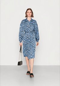 Vestido azul com padrão, mangas longas, cintura ajustada e colarinho com botões. Usado com sandálias pretas rasas e uma pequena bolsa preta.