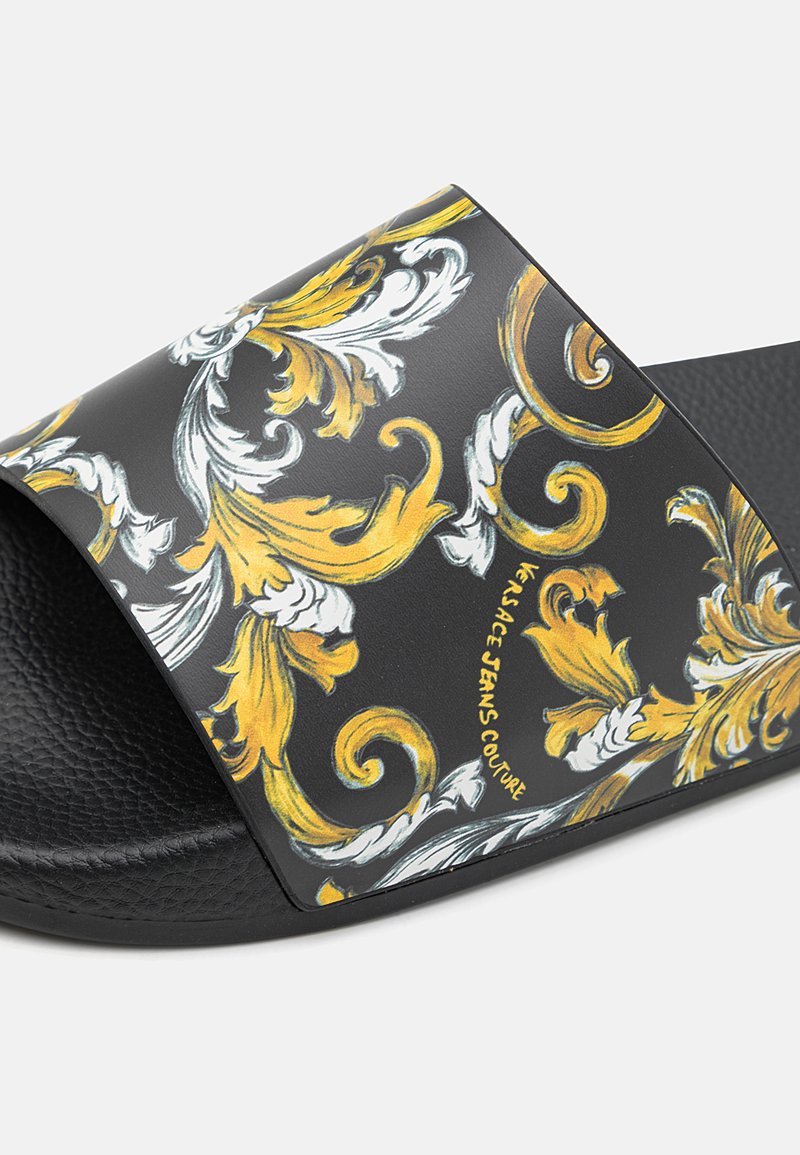 Sandales noires à plateforme avec un motif floral en or et blanc sur la bride. Texte : "Versace Jeans Couture." Semelle noire texturée.
