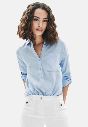 Femme aux cheveux ondulés mi-longs portant une chemise bleu clair à boutons avec les manches retroussées et un pantalon blanc, debout les mains dans les poches.