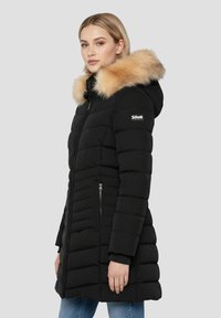 Veste matelassée noire avec capuche en fausse fourrure, poches avant zippées et texture matelassée. Présente un logo sur l'épaule gauche.
