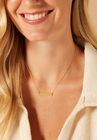 Gouden ketting met een rechthoekige hanger, een dunne ketting en een gladde afwerking, gedragen tegen een lichtgetextureerde stof. Huidskleur is neutraal.