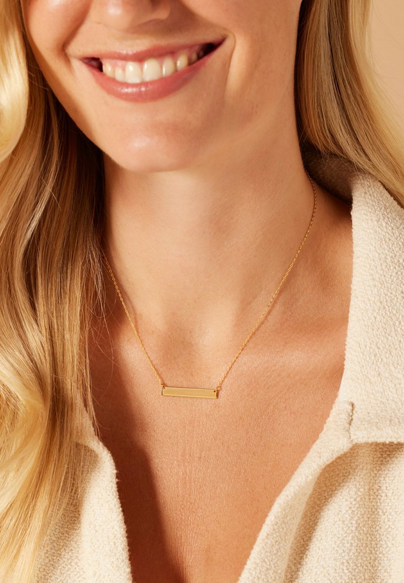 Gouden ketting met een rechthoekige hanger, een dunne ketting en een gladde afwerking, gedragen tegen een lichtgetextureerde stof. Huidskleur is neutraal.