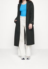 Abrigo largo de color verde oscuro con cuello muesca, llevado sobre una camiseta azul con relieve, acompañado de pantalones de pierna ancha blancos y zapatillas.