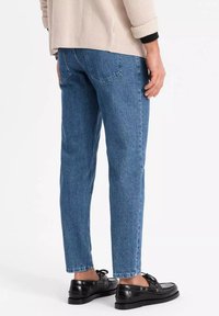 Persona vistiendo jeans de mezclilla azules, suéter marrón claro y mocasines de cuero negros con cordones, de pie contra un fondo blanco liso.