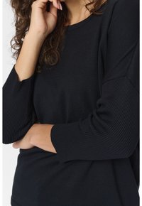 Femme aux cheveux bouclés portant un pull noir côtelé à manches longues, une main reposant près du visage et l'autre croisée sous le coude.