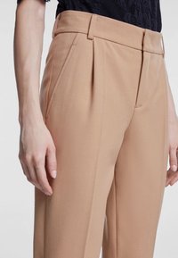 Beige skræddersyede bukser med høj talje, folder og sidelommer, lavet af et glat stof. Modellens hånd hviler på buksebenet.