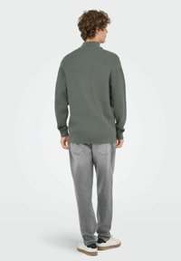 Suéter de punto de manga larga en verde oliva con cuello doblado, combinado con pantalones tapered en gris claro y zapatillas blancas con detalles en negro.