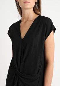 Svart klänning med djup V-ringning, korta ärmar med kapprar och texturerad tyg. Har en knuten detalj vid midjan för extra dimension.