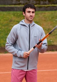 Ljusgrå zip-up hoodie med en svart logotyp, bärs över en vit skjorta; ihop med rosa shorts. Personen håller ett tennisracket.