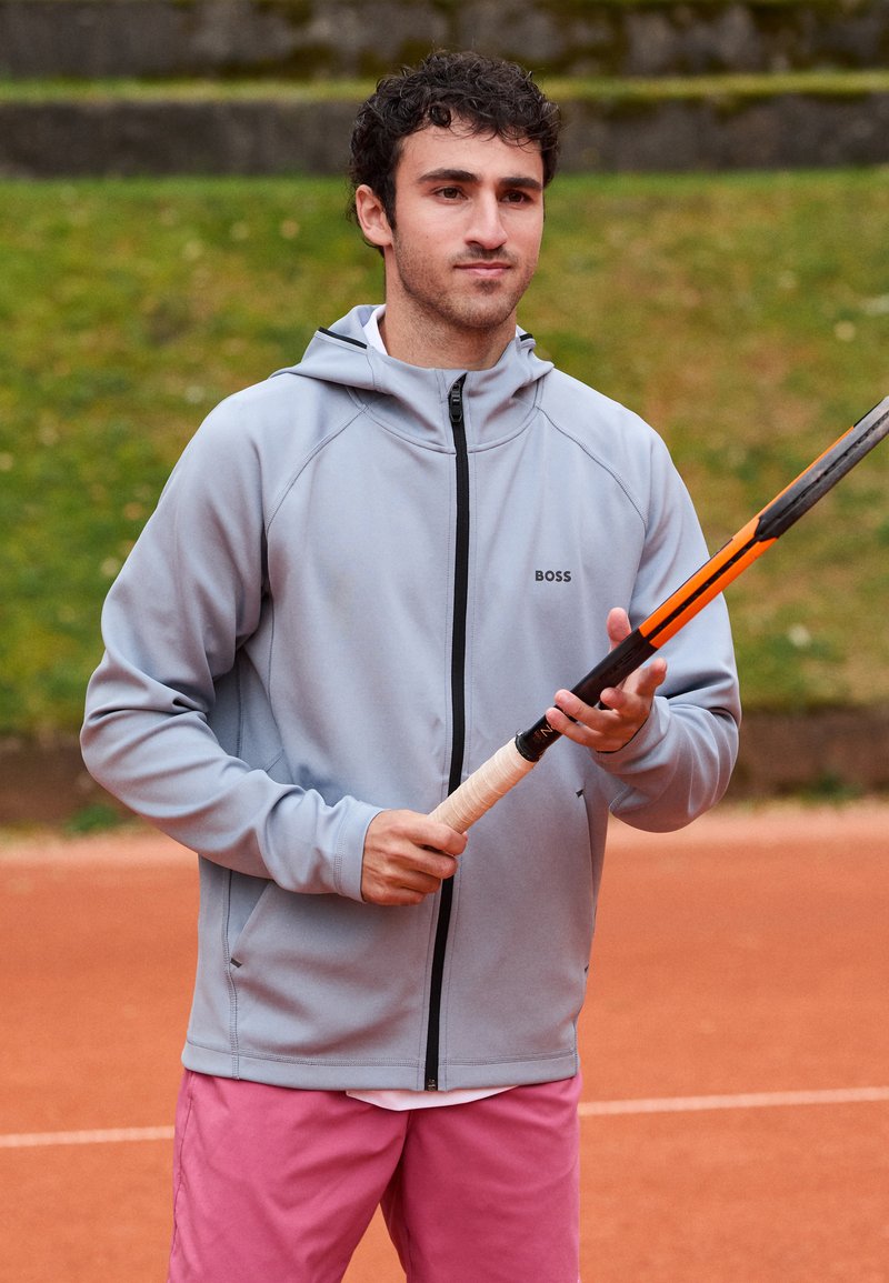Ljusgrå zip-up hoodie med en svart logotyp, bärs över en vit skjorta; ihop med rosa shorts. Personen håller ett tennisracket.