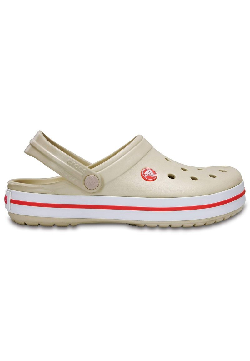 Beige Crocs with side holes, a back strap, and white midsole featuring a red stripe. Rubber material with textured surfaces.