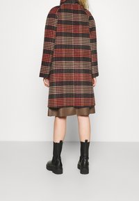 Persona vistiendo un abrigo de cuadros marrón y rojo, una falda marrón y botas negras de tobillo, de pie contra un fondo claro liso.