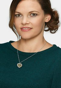 Sølv kæderegister med en hjerteformet vedhæng med et indviklet åbenværksdesign, båret over en mørkegrøn, struktureret sweater.
