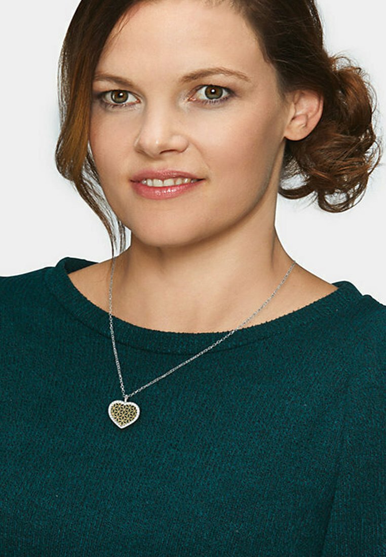 Sølv kæderegister med en hjerteformet vedhæng med et indviklet åbenværksdesign, båret over en mørkegrøn, struktureret sweater.