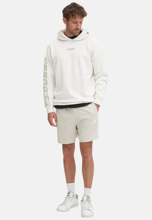 Felpa bianca con branding "GUESS" sulla parte anteriore e sulla manica, abbinata a pantaloni corti beige con un logo, e sneakers bianche. Design e materiali semplici.