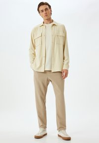 Lys beige skjorte med knapper og to brystlommer, lagd over en cremet t-shirt, parret med beige tapered bukser og hvide sneakers.