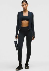 Fekete sportos öltözet, beleértve egy cipzáras kabátot, sportmelltartót és magas derekú leggingset. Egy sötét vízpalackot tart kezében, rajta logóval.