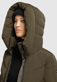 Dik olijfgroene jacket met een grote capuchon en een contrasterende zwarte rits. Heeft een geborduurd patroon en wordt gedragen over een zwart shirt.