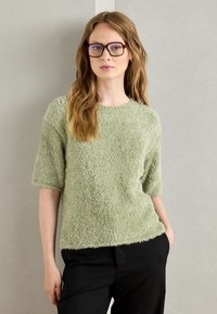 Light green fluffy textured sweater with short sleeves, paired with black trousers. Model has wavy hair and is wearing glasses.
