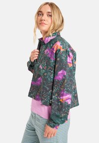Bunte Jacke mit floralem und pflanzlichem Muster, mit Kragen, Fronttasche und lockerer Passform. Der Stoff wirkt strukturiert.