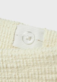 Krämfärgad ribbad tyg med texturerad väv, med ett vitt vävt band och en genomskinlig knapp fäst. Mjuk och plyschig utseende.