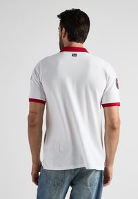 Mężczyzna w białej koszulce polo z czerwonym kołnierzykiem i wykończeniami rękawów, stojący tyłem do kamery, widoczne niebieskie dżinsy, tatuaż na lewym ramieniu.