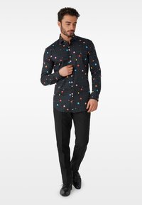 Schwarze Hemdbluse mit einem Pac-Man-Spielmuster in bunten Farbtönen. Kombiniert mit schwarzen Hosen und Schuhen, die ein schlankes Design aufweisen.