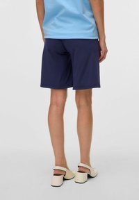 Jūras spēku šorti, izgatavoti no mīksta auduma, līdz ceļgaliem, ar vienkāršu dizainu un bez redzamām rakstu. Modelis valkā krēmkrāsas blokveida papēžus.