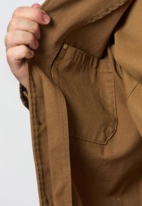 Main tenant une veste marron ouverte montrant une poche intérieure avec une fermeture à bouton pression.