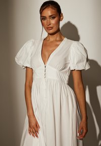 Weißes Baumwollkleid mit puffigen Ärmeln, V-Ausschnitt, Vorderknöpfen und ausgestelltem Rock. Die sanfte Textur und das fließende Design schaffen eine entspannte Silhouette.