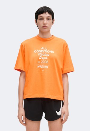 Persona con cabello oscuro y corto lleva una camiseta naranja brillante con texto blanco y pantalones cortos negros de Nike sobre un fondo claro liso.