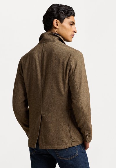 Braune Wolljacke mit hohem Kragen, geknöpften Manschetten und einem dezenten Fischgrätenmuster. Der Rückenschlitz verleiht Struktur. Kombiniert mit dunklen Jeans.