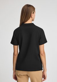 Polo-shirt noir avec un col, des manches courtes et une coupe droite. Confectionné en tissu lisse, il dispose de fentes latérales pour un confort de mouvement.