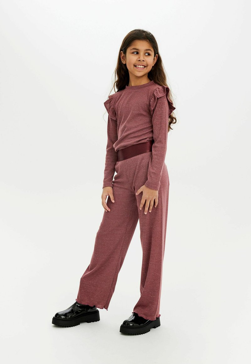 Vinröd tvådelad outfit med volangärmar och matchande vida byxor. Slätt, texturerat tyg; parat med svarta klumpiga skor.