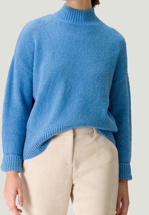 Pull bleu à col montant texturé avec poignets retroussés, associé à un pantalon beige taille haute porté par une personne debout.