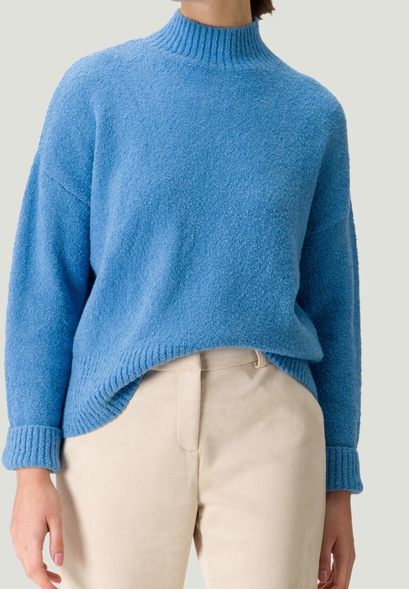 Pull bleu à col montant texturé avec poignets retroussés, associé à un pantalon beige taille haute porté par une personne debout.